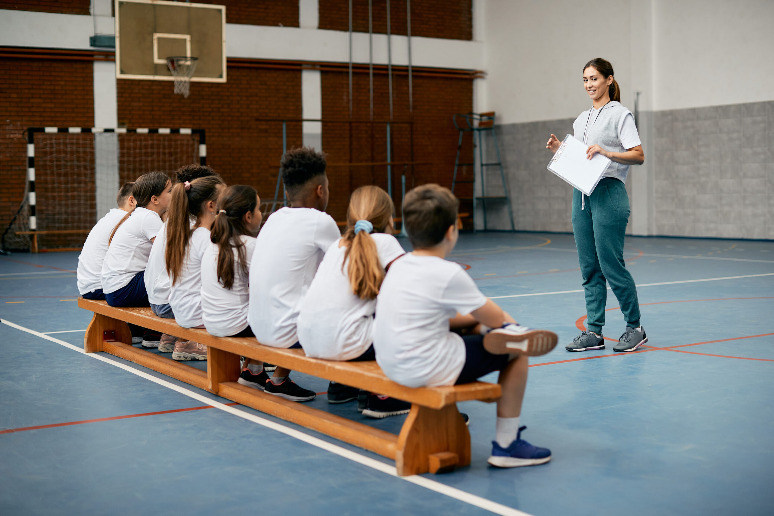Young female coach having PE class with group of elementary students at school gym.