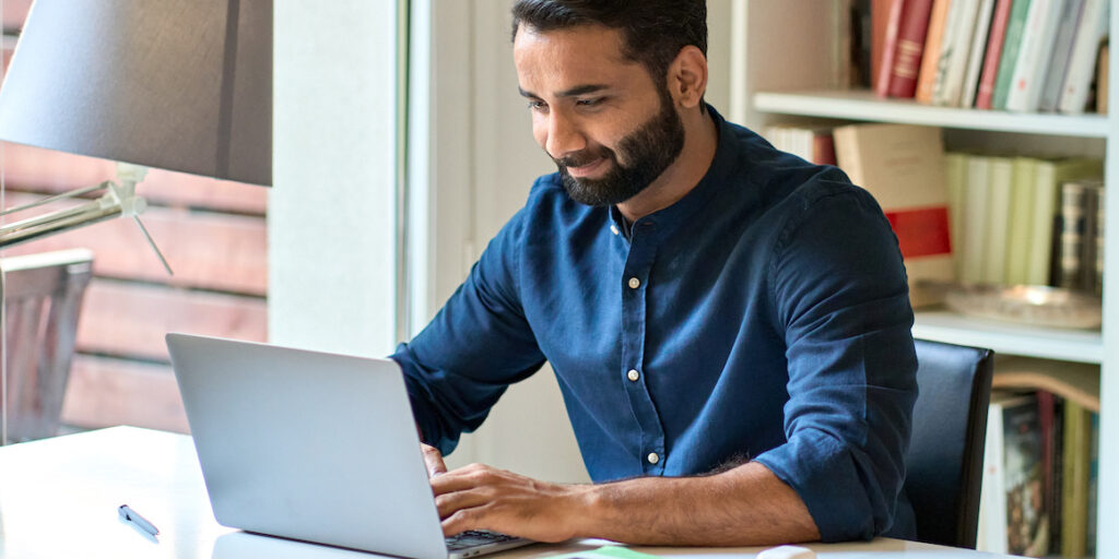 A man sitting at a library desk, taking notes, and looking at a laptop