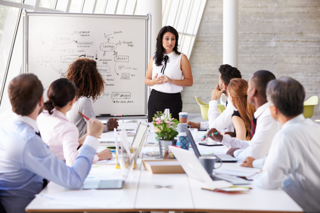 A professional walks her team through an explanation of organizational change management. She stands next to a whiteboard in front of a group of businesspeople.