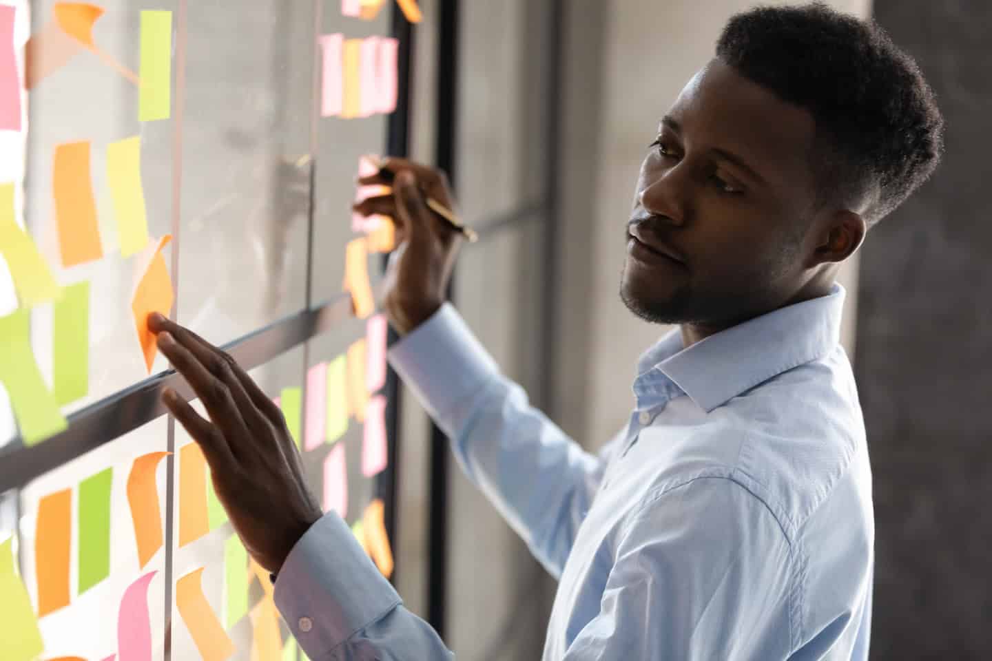 Serious african businessmen managing project tasks on sticky notes, focused male professional coach teacher write creative business idea on post it stickers planning corporate strategy on glass board