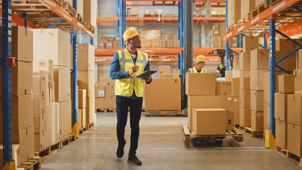 A worker wearing a hard hat and safety vest while walking through a warehouse stocked with boxes.