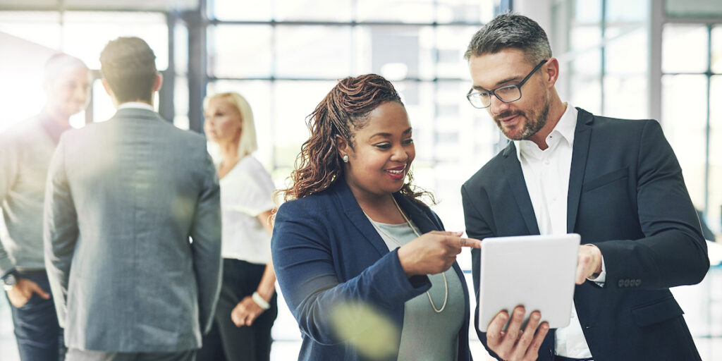 Two business professionals looking at information on a tablet together. A group of professionals is chatting behind them.