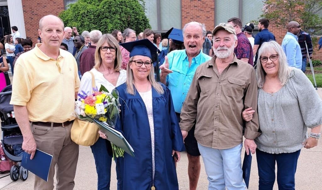 A young woman in a blue cap and gown stands proudly outside the MVNU campus, holding a bouquet of flowers and her college degree. Surrounded by her immediate and extended family, she smiles joyfully. Other graduates and their families are visible in the background, celebrating their achievements together.