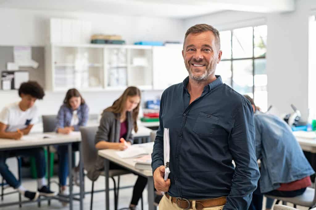 A mature teacher, conveying a sense of professionalism and satisfaction, stands confidently in his classroom after advancing his teaching career through MVNU’s online Master’s in Education program