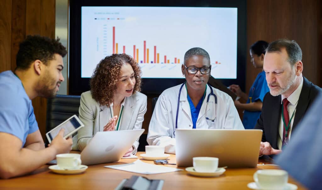 Healthcare professionals and administrators in a hospital boardroom collaborate during a leadership meeting, reviewing charts and data on digital devices. This image reflects the kind of real-world, strategic healthcare leadership roles that MVNU’s online MBA in Healthcare Administration prepares students to step into.