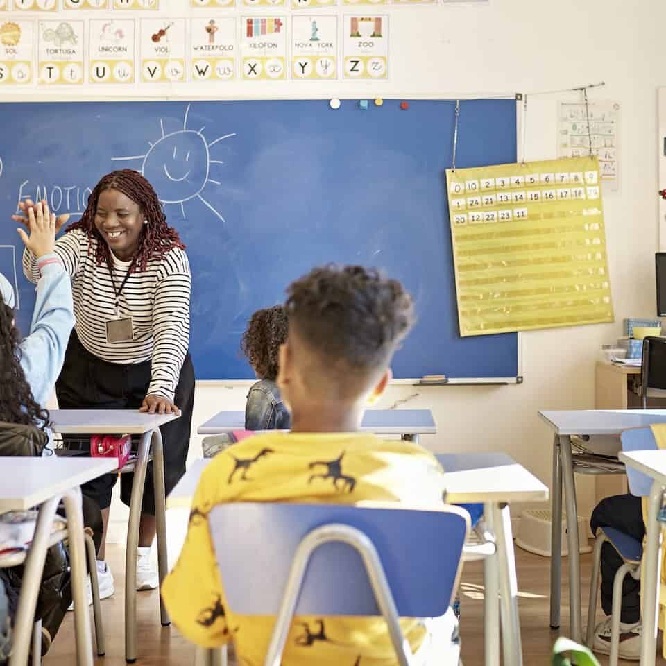 View from behind classmates seated at child-size desks of smiling mid 40s female instructor supporting student with encouraging gesture.