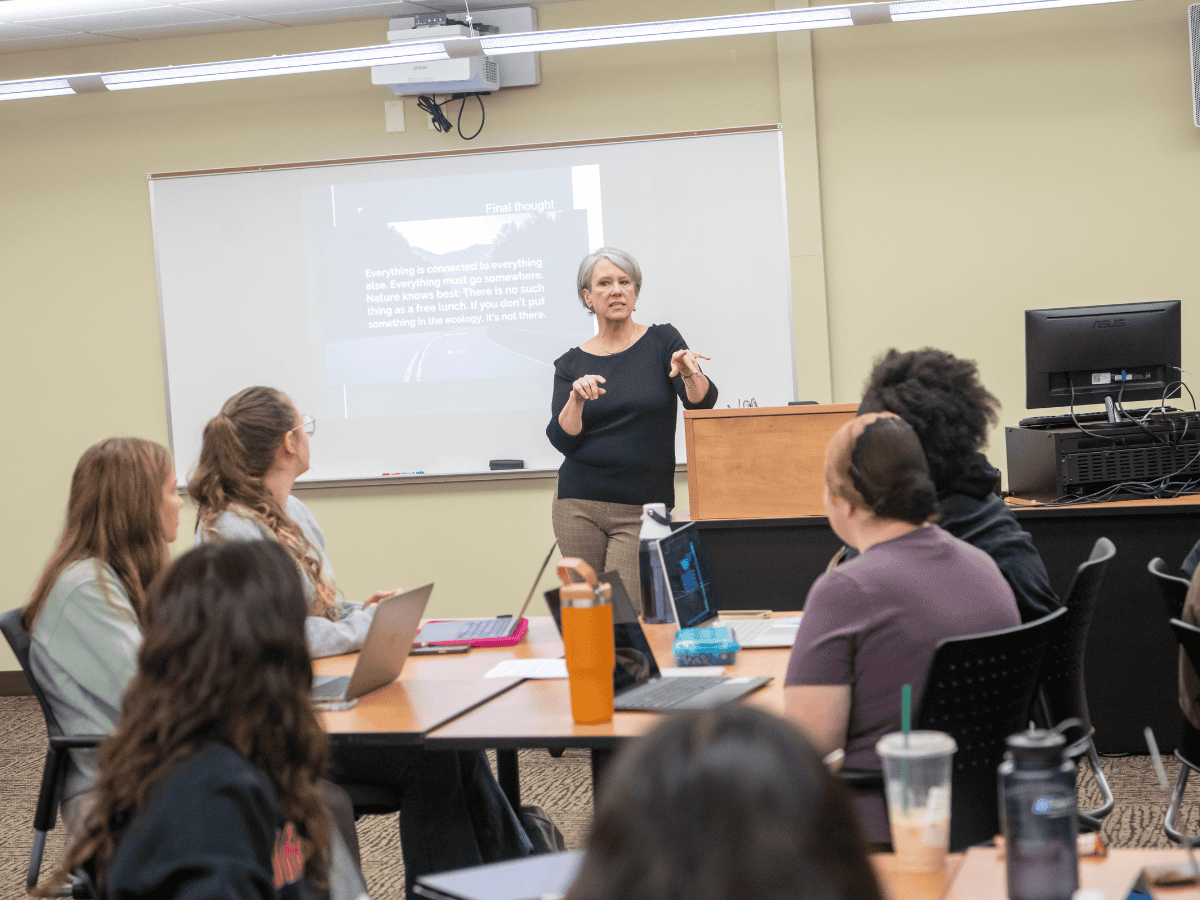 Classroom lecture with a female instructor presenting to students using laptops.