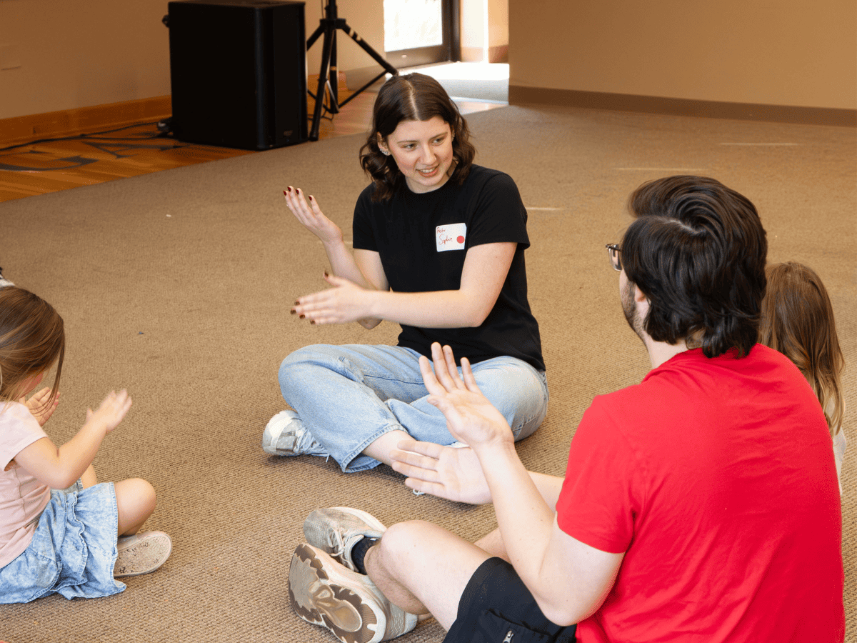 A young woman and a man engage children in a pastoral ministry activity, sitting on the carpeted flo.