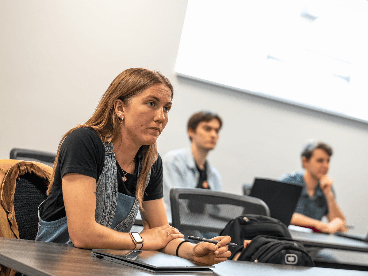 Woman attentively listening during a classroom lecture with students in the background.