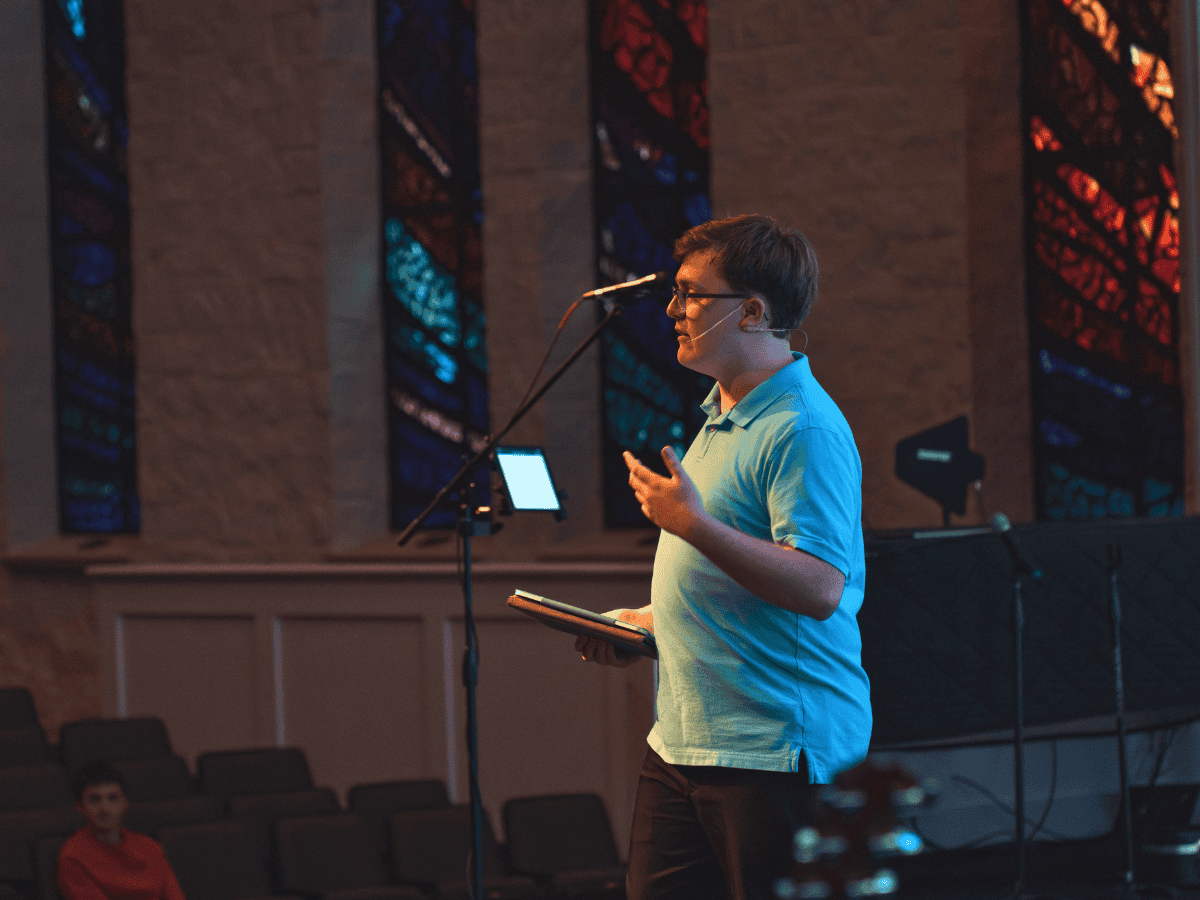 Young man delivering a speech or sermon in a church setting with stained glass windows.