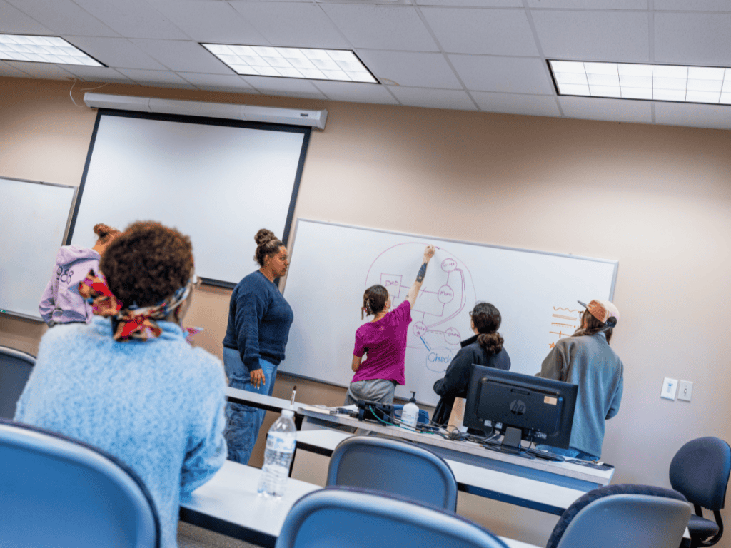 A woman writes on a whiteboard during a classroom presentation with students seated at desks.