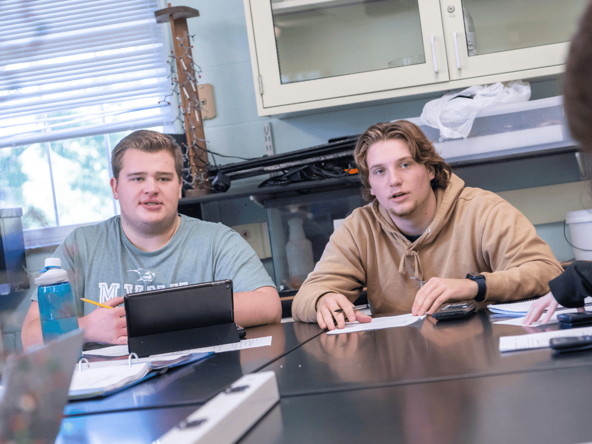 Two male students sitting at a table with notebooks and tablets, engaged in a discussion in a classr.