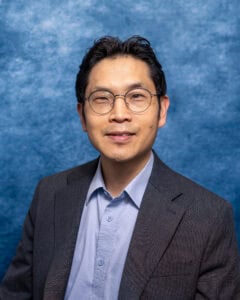 Senior professional man in a suit with glasses, smiling, against a blue textured background.
