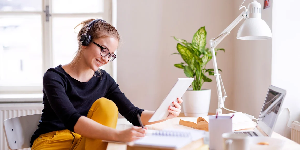 Smiling adult student wearing headphones studies at home, using a tablet and laptop while taking notes—representing the flexibility and convenience of earning a degree online.