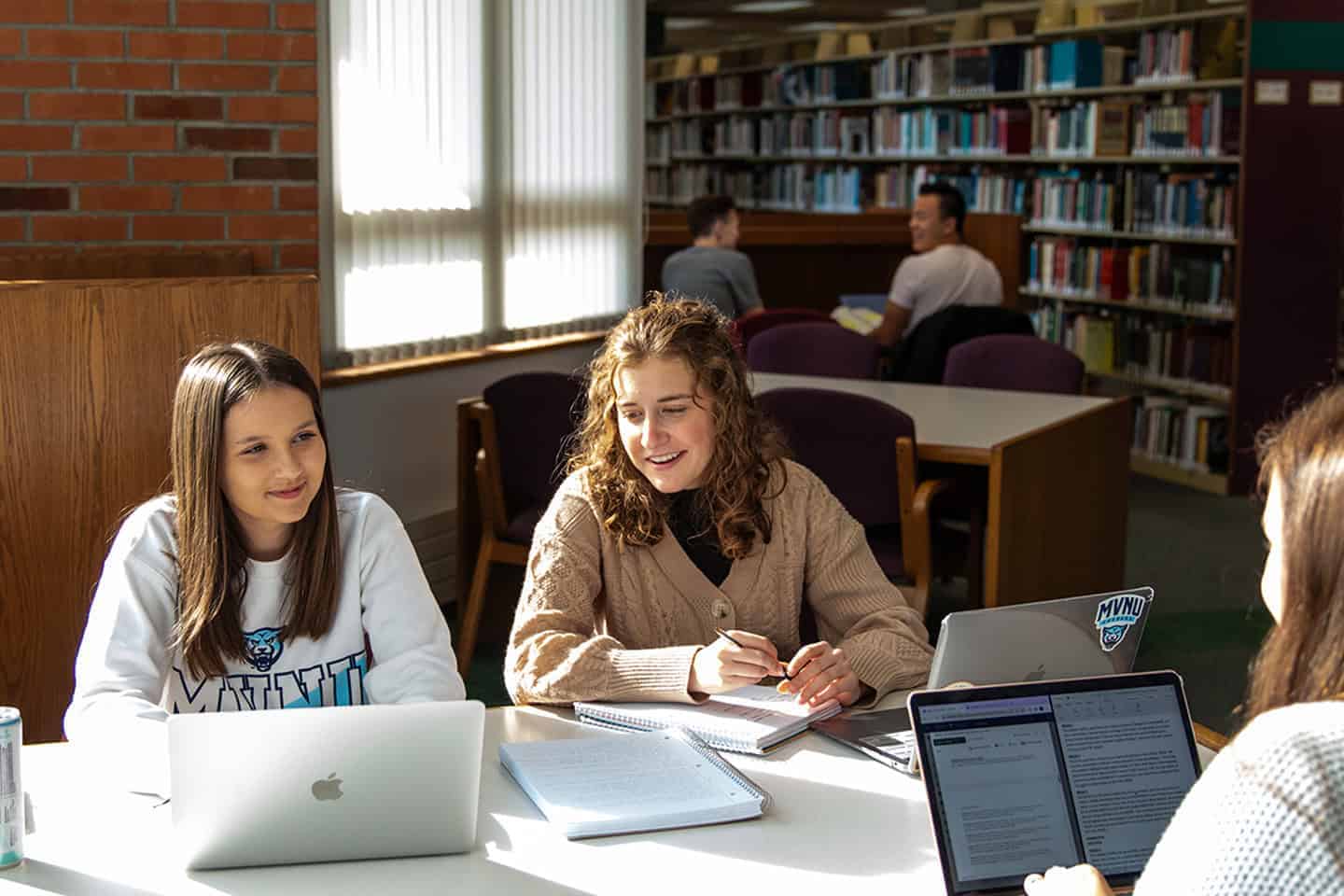 Students studying and collaborating at a library table with laptops and books.