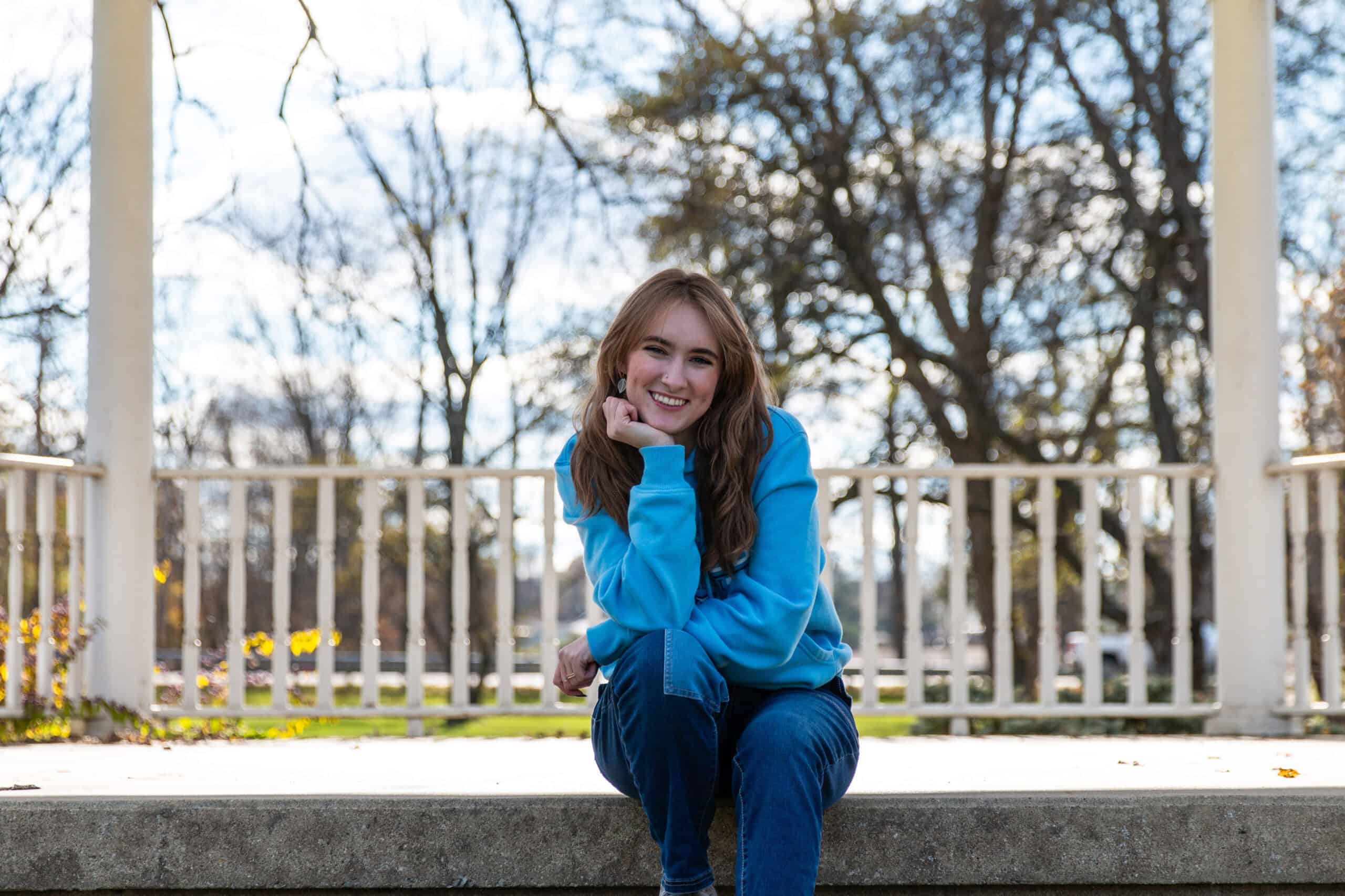 MVNUADM-6831 A young woman with long brown hair wearing a light blue hoodie and jeans sits on a stone ledge, smiling brightly with her head resting on her hand. She is outdoors in a park-like setting with bare trees and a white railing behind her, under a partly cloudy sky.