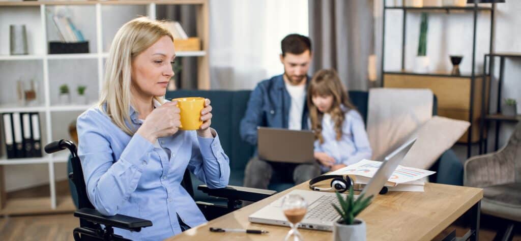 Woman in wheelchair, while using a laptop at home to attend an online MVNU MBA course, with her husband and child playing in the background