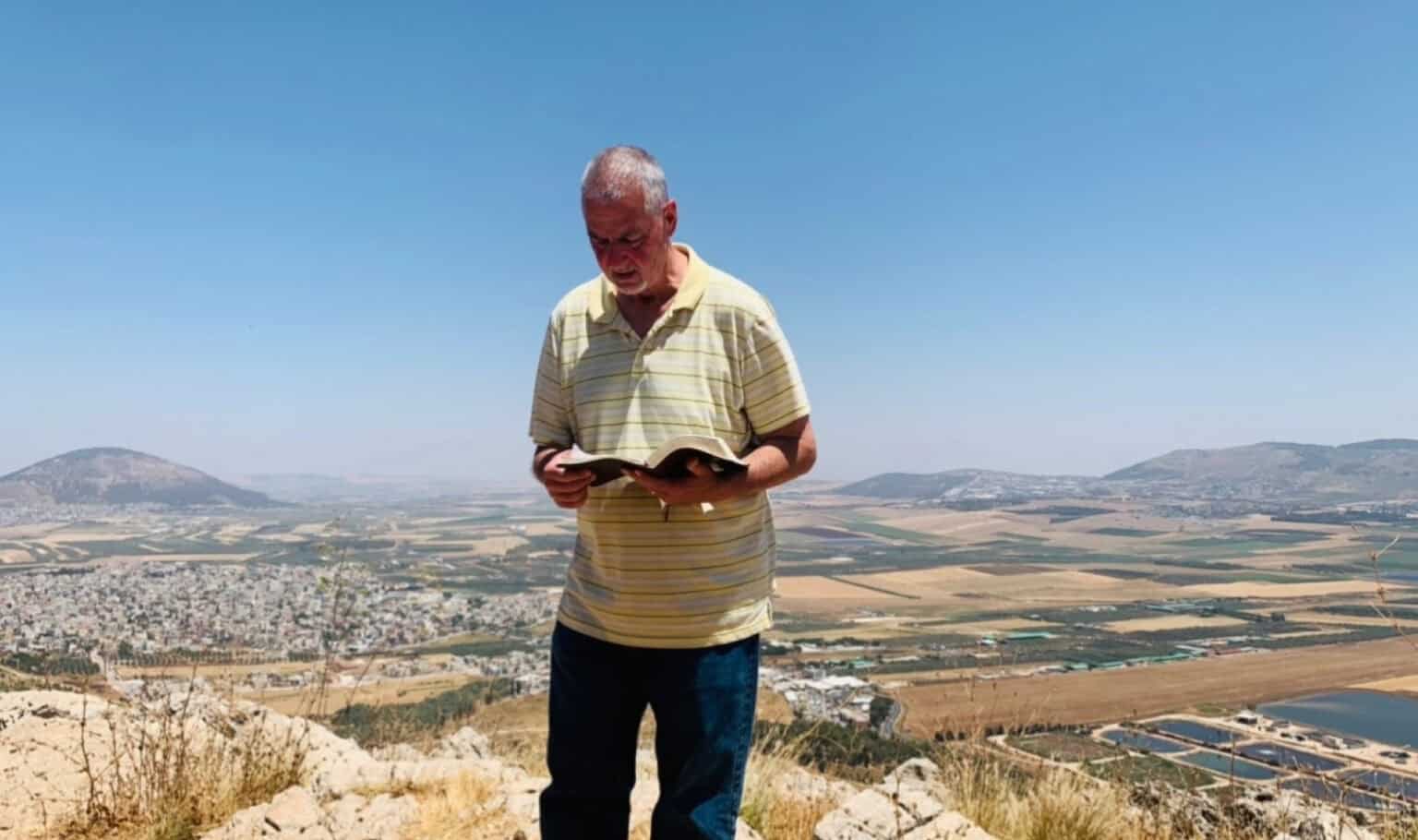 Dr. Mike VanZant, MVNU professor of biblical literature, stands outdoors on a mountaintop in Israel, reading from the Bible with a scenic valley landscape behind him.