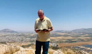 Dr. Mike VanZant, MVNU professor of biblical literature, stands outdoors on a mountaintop in Israel, reading from the Bible with a scenic valley landscape behind him.