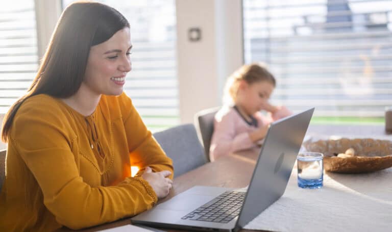 Smiling woman attends an MVNU online class on her laptop at a kitchen table, while a young child plays in the background.