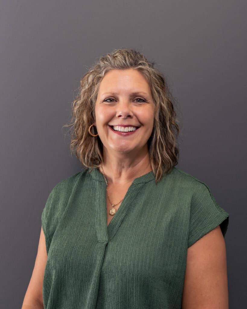 A smiling woman with curly blonde hair wearing a green blouse against a gray background.
