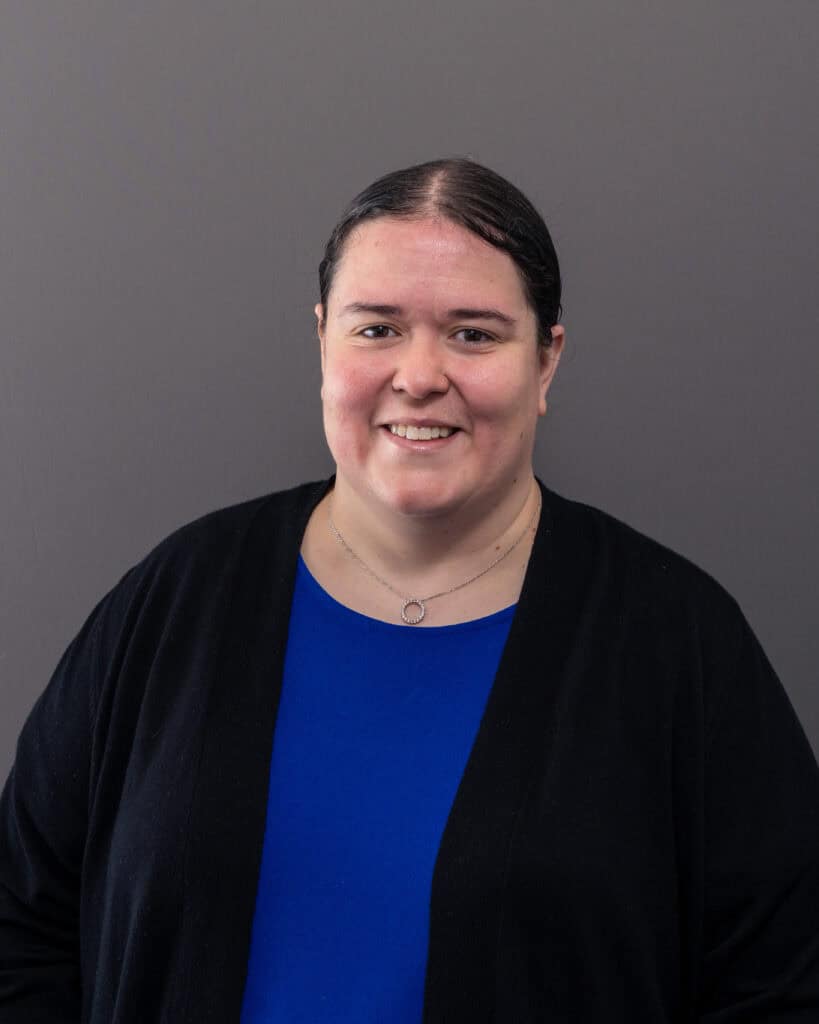 A woman smiling in front of a gray background, wearing a blue top and black cardigan.