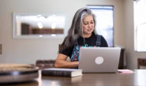 An adult student studies at home using her laptop, representing the flexibility of MVNU Online programs. She sits at a dining table with study materials nearby, focused and engaged in her coursework in a bright, comfortable home environment.