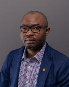 Senior professional man wearing glasses and a blue blazer, looking serious against a gray background.
