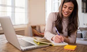 A smiling adult woman sits at a table with a laptop open and notebook in front of her, taking notes for an online class. The bright, home-like setting and natural light convey the flexibility of earning a business degree online through MVNU’s adult and graduate programs.