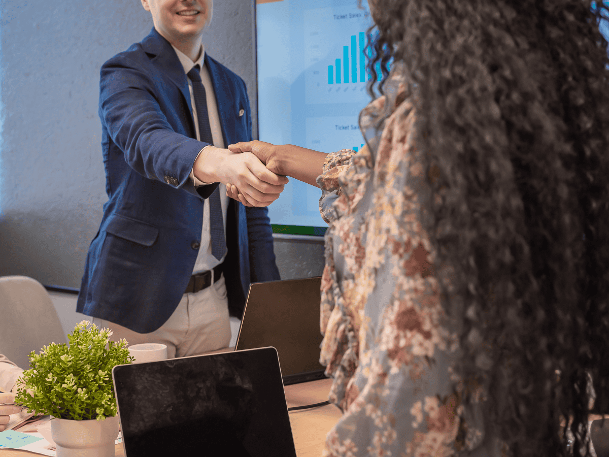 Handshake between two professionals in a business meeting, with a presentation screen showing graphs.