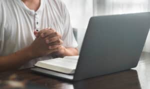 Adult student sitting at a desk with a Bible open and hands folded in prayer beside a laptop, representing faith integrated with online learning.