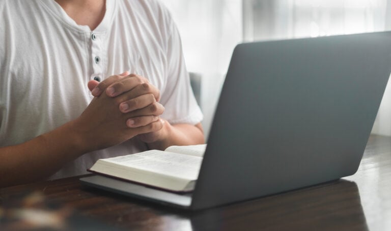 Adult student sitting at a desk with a Bible open and hands folded in prayer beside a laptop, representing faith integrated with online learning.