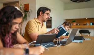 A parent studying with a laptop and notebook alongside two children at a table, representing how MVNU Online students balance schoolwork with family life.
