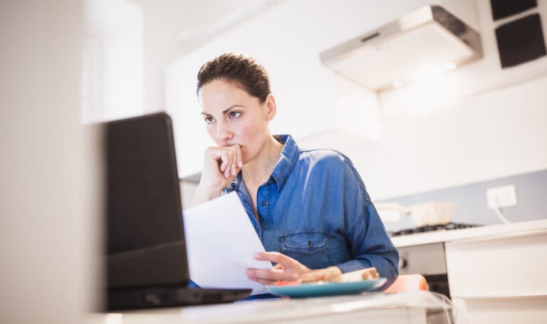An adult woman studying at home with a laptop and paperwork in her kitchen, representing the real-life balance many MVNU Online students manage while returning to college.