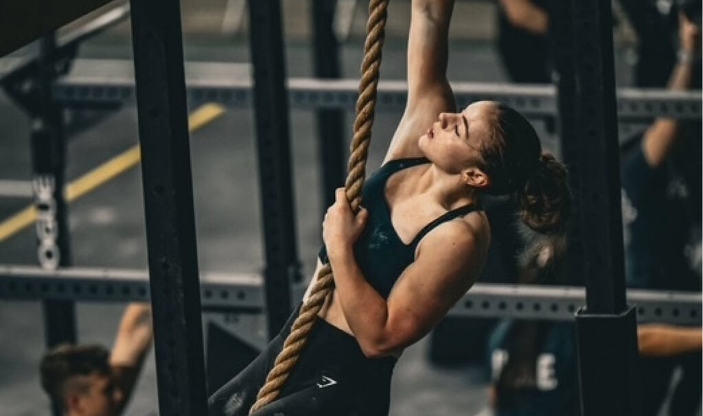 Elsie L. trains in a CrossFit gym, climbing a rope during an intense workout session while pursuing her associate degree through MVNU Online.