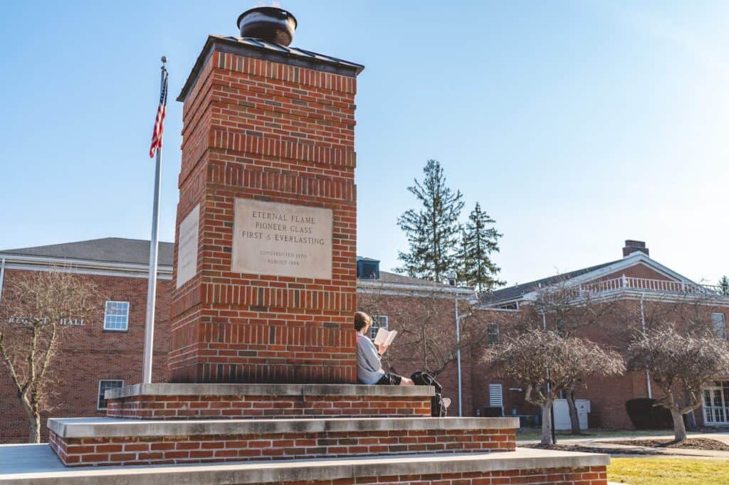 A woman sitting on a brick monument reading a book outside a university building.