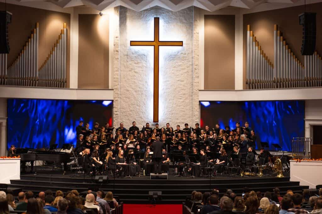 A church choir performing on stage with a conductor in front, in a modern church interior with a lar.