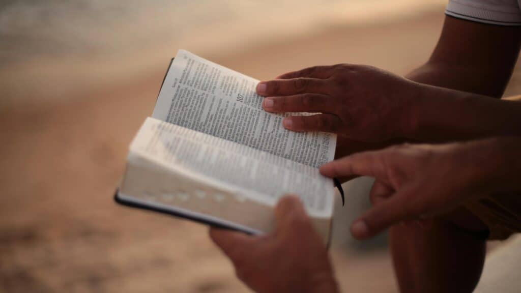 A person reading a book outdoors on a sandy surface.