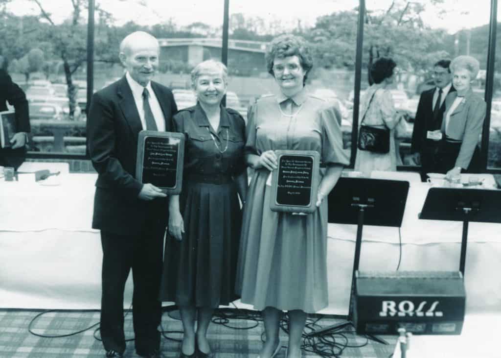 Image of Dr. Lora “Doc” Donoho with two colleagues holding plaques at an event.