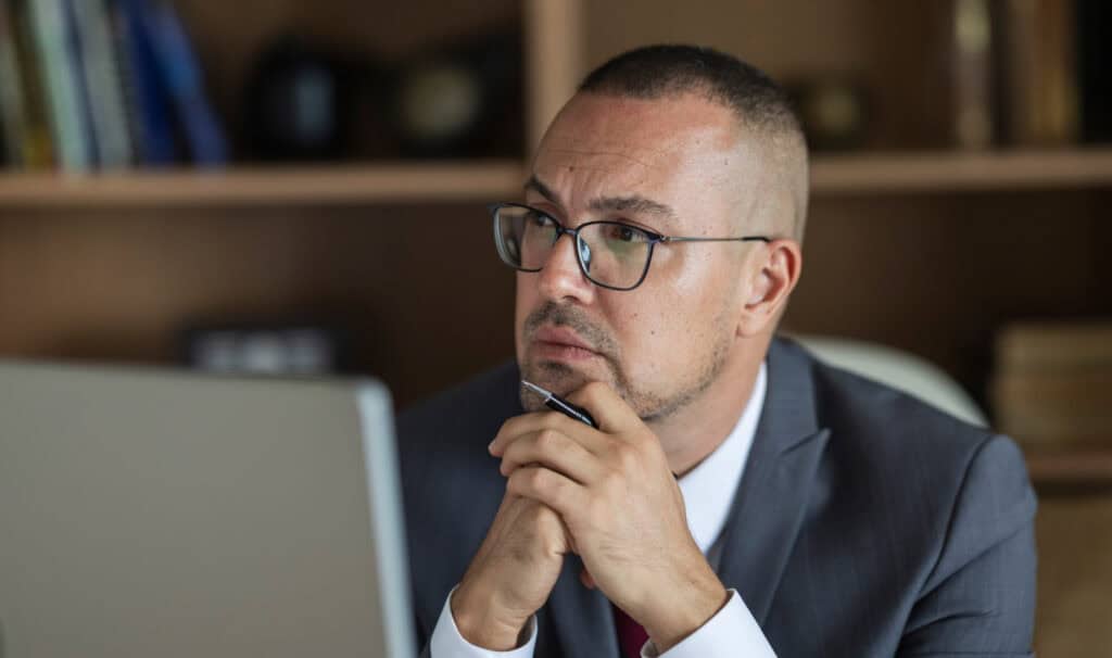 Businessman contemplating work at his desk with a laptop and bookshelf in the background.