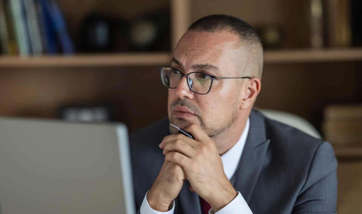 Businessman contemplating work at his desk with a laptop and bookshelf in the background.
