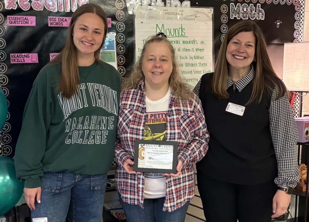 Three women smiling, one holding a certificate, in a classroom setting.