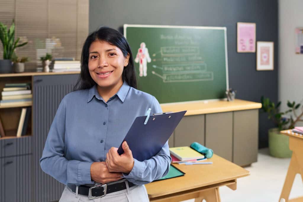 A woman teacher smiling in classroom holding a clipboard.
