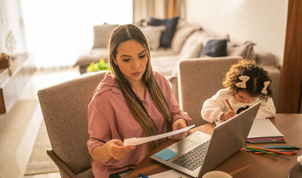 A mother studies at a dining room table with a laptop and documents while her young daughter colors nearby, representing the real-life demands online students balance at MVNU Online.