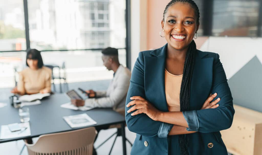 A confident professional woman stands with arms crossed and smiling in a modern office, with colleagues working at a conference table behind her, representing the leadership presence and career advancement MVNU Online's Master of Organizational Leadership program develops.