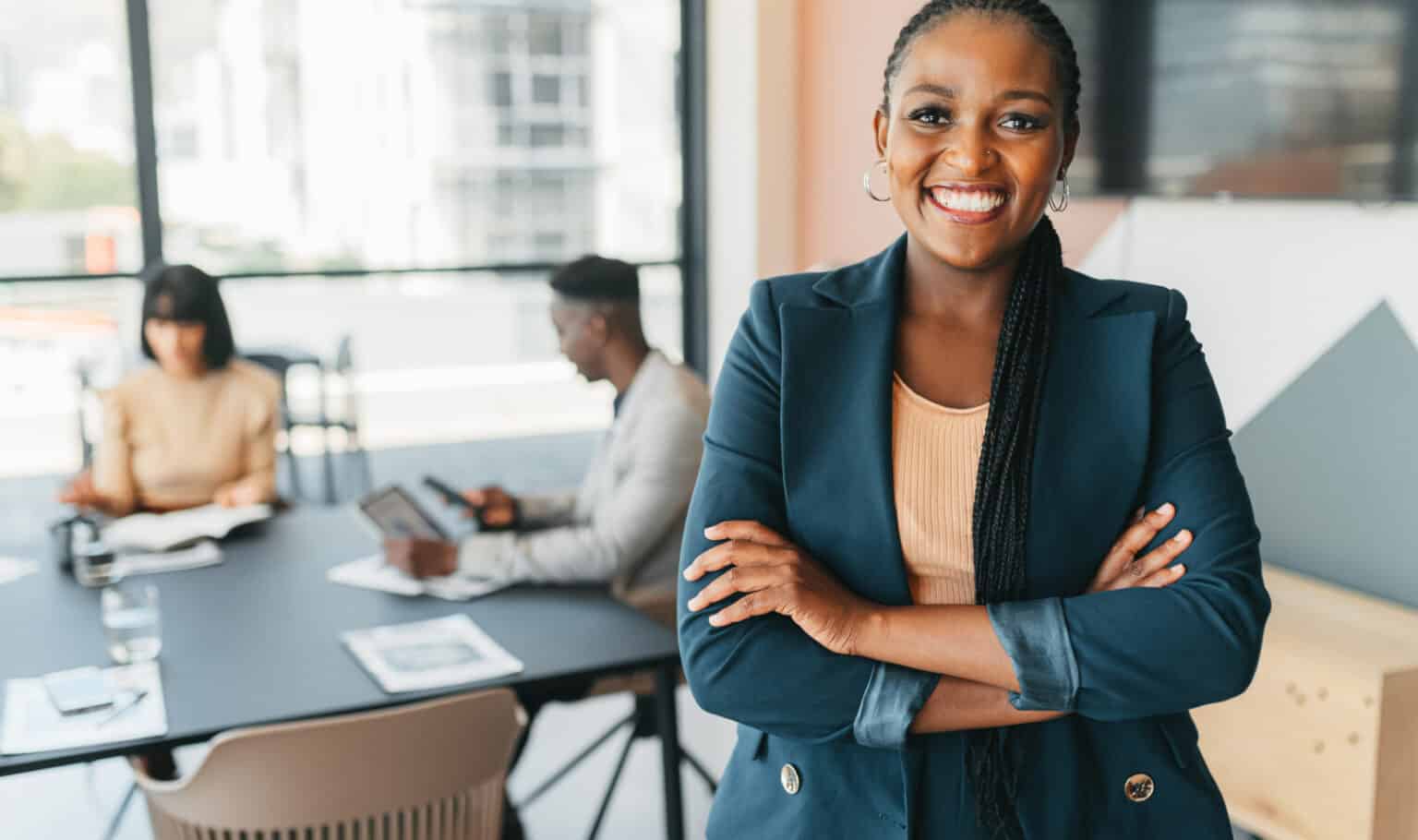 A confident professional woman stands with arms crossed and smiling in a modern office, with colleagues working at a conference table behind her, representing the leadership presence and career advancement MVNU Online's Master of Organizational Leadership program develops.
