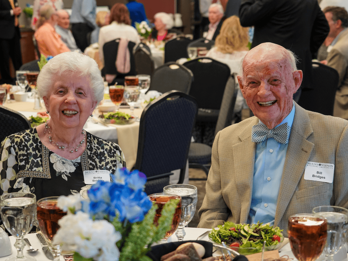 Smiling elderly couple at a formal event with tables and floral centerpieces.