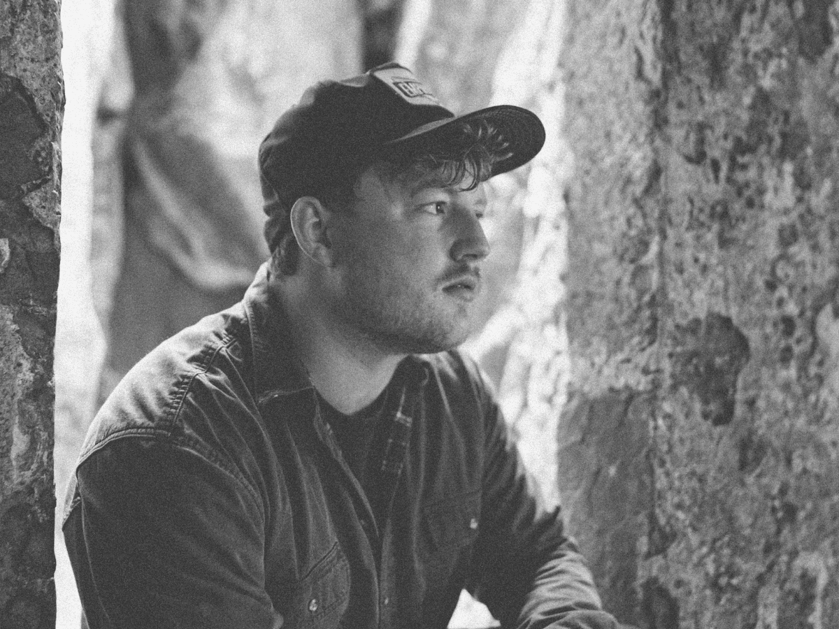 Young man in outdoor setting with a cap and jacket, looking thoughtfully into the distance.