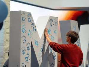 Student interacts with large 'M' letter decorated with blue handprints.
