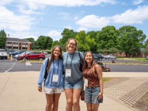Three smiling students at orientation on a sunny day outside campus.